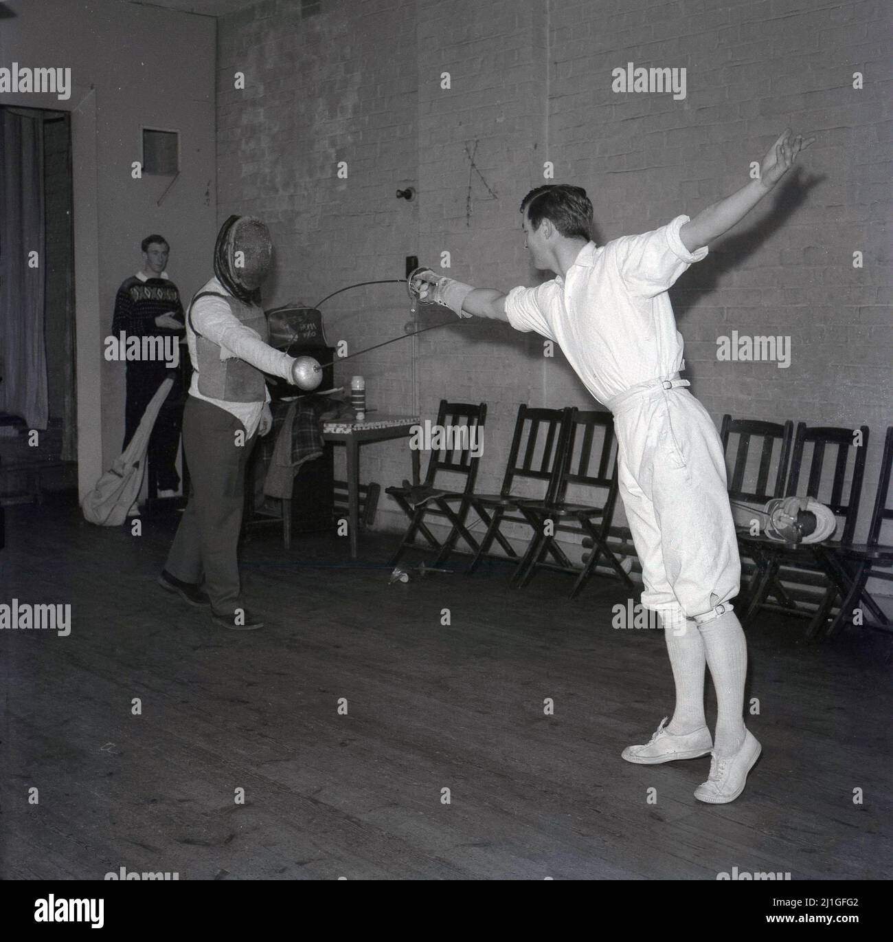 1960s, storico, classe di scherma, all'interno di una sala, uno studente maschile che indossa tradizionali breches scherma, praticando con un istruttore o insegnante di scherma, Università di Oxford, Inghilterra, Regno Unito. Stanno usando le epee, un'arma di thusting, un po'più pesante di un foglio, un'altra arma di scherma. L'istruttore indossa una maschera per il viso e una leggera giacca protettiva nota come zoppo. Uno sport di combattimento elegante ma foscialmente impegnativo, la scherma è uno dei soli cinque sport da avere in ogni moderno Giochi Olimpici. Foto Stock