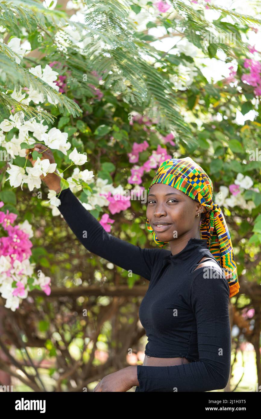 Deliziosa ragazza africana giovane, con un archetto colorato, che raccoglie fiori da un albero in un giardino tropicale. Foto Stock