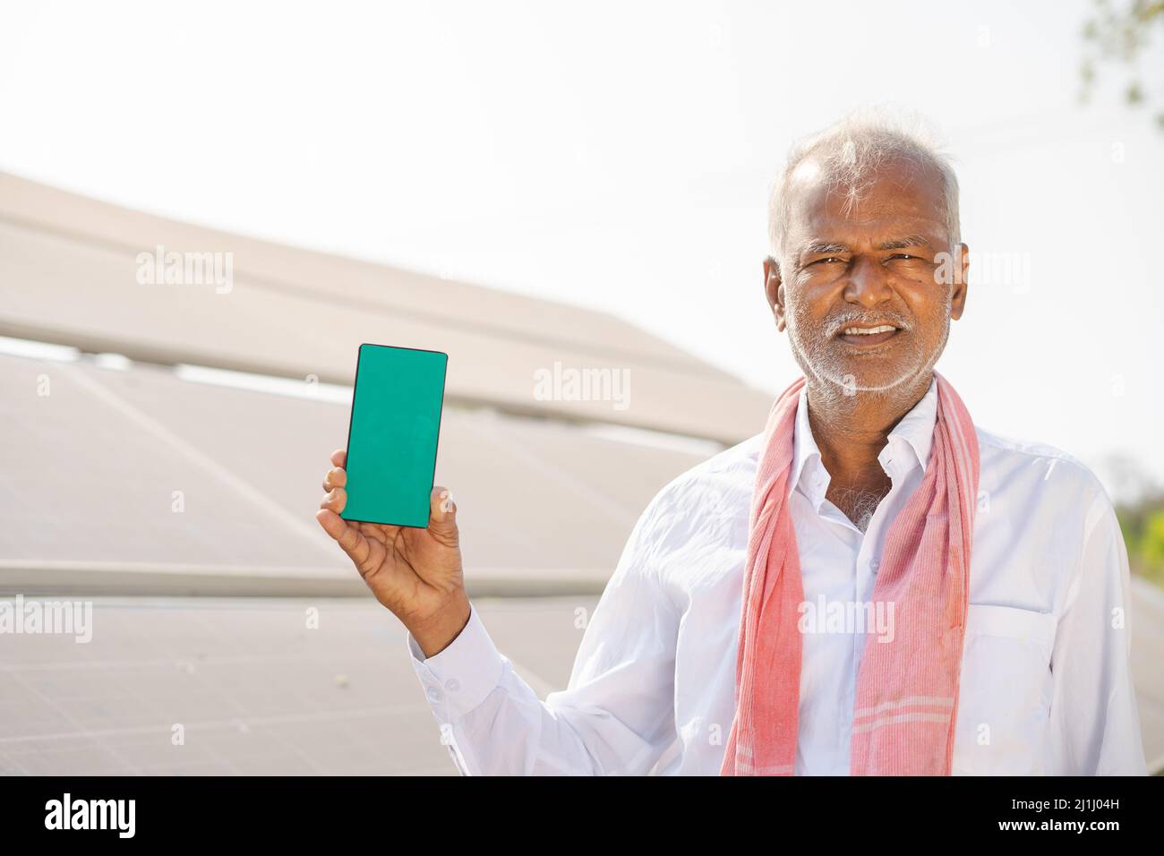 un agricoltore sorridente che mostra il telefono cellulare con schermo verde puntando il dito mentre si trova di fronte al pannello solare in campagna - concetto di pubblicità app Foto Stock