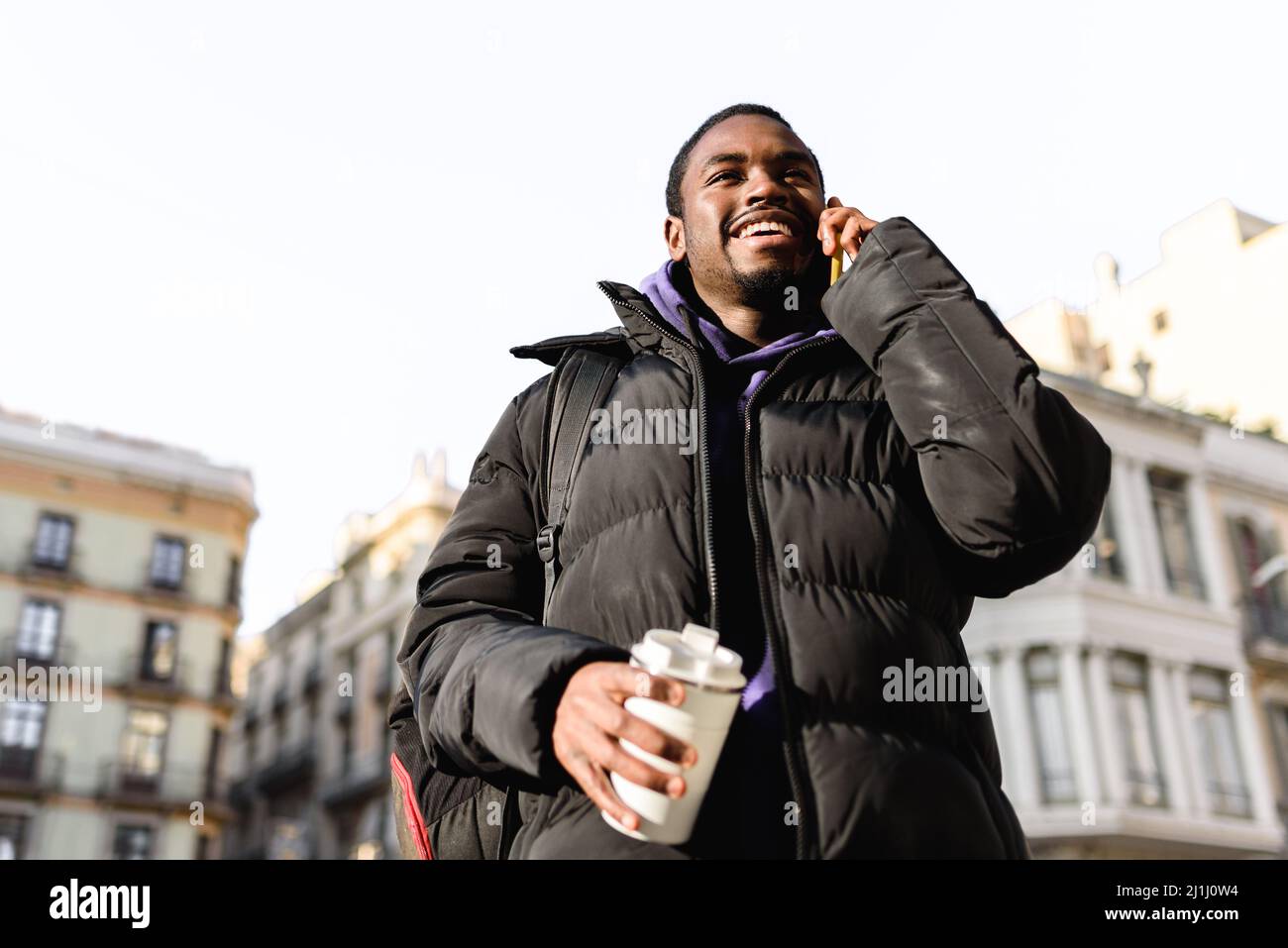 Da sotto il maschio nero felice in outerwear con zero rifiuti takeaway tazza di bevanda calda. Sorride allegro e parla al telefono cellulare su sfondo sfocato sulla strada della città in giorno di sole Foto Stock