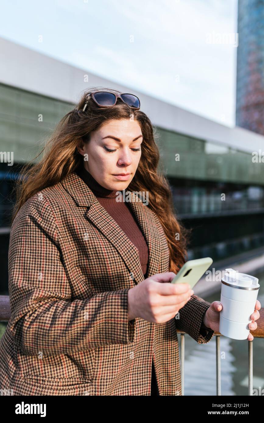 Donna in cappotto a scacchi con zero tazzina di takeaway caffè navigazione cellulare mentre in piedi sul ponte in città di giorno Foto Stock