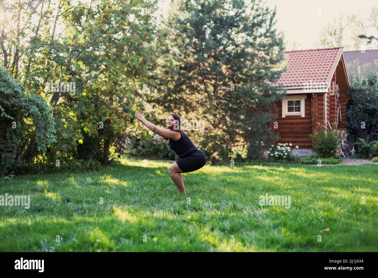 Giovane donna grasso guardando la macchina fotografica facendo yoga squatting con mani in su posizione sul cortile di cottage con casa di legno e alberi in background. Corpo Foto Stock