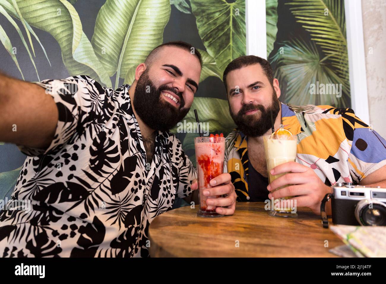 Felice giovane coppia gay in estate vestiti prendendo un autoritratto insieme mentre si tiene un cocktail durante la loro vacanza. Foto Stock