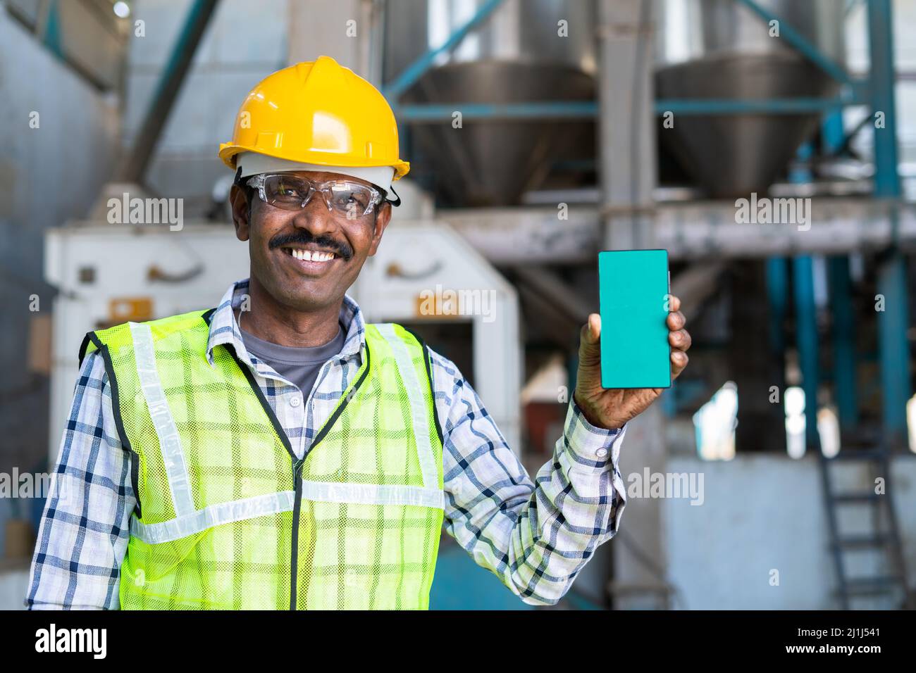 Sorridente lavoratore industriale che mostra il telefono cellulare con schermo verde mentre guarda la macchina fotografica - concetto di servizio di prenotazione, pubblicità e promozione. Foto Stock