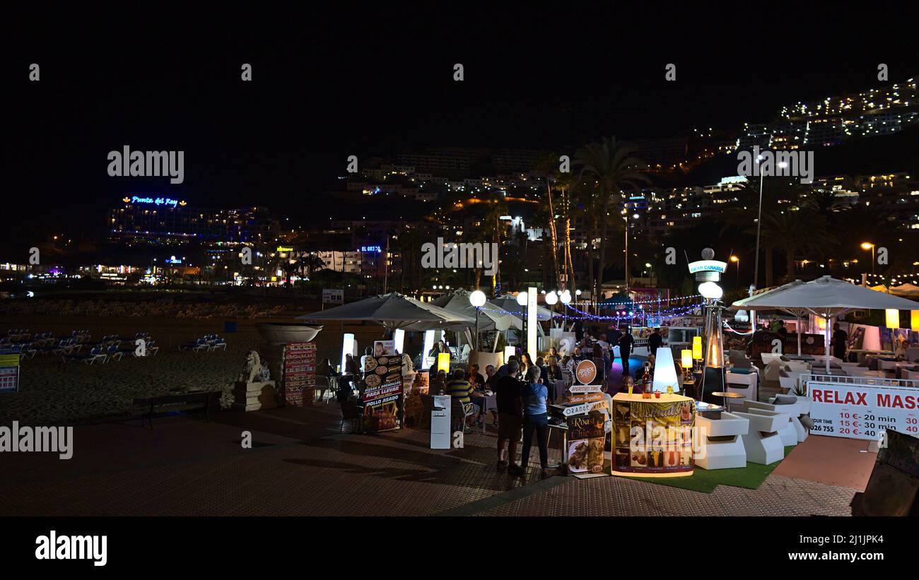 I turisti si godono la cena al ristorante vicino alla spiaggia di Puerto Rico, Gran Canaria, Spagna in serata con edifici illuminati dell'hotel. Foto Stock