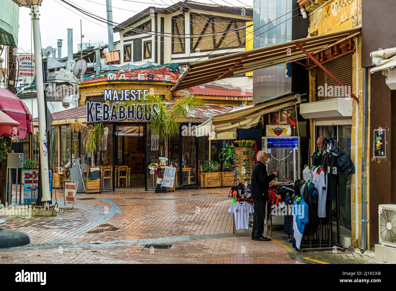 La zona pedonale del centro storico di Lefkoşa (Nicosia), Repubblica Turca di Cipro del Nord (RTNC), passa qui davanti al ristorante “Majeste Kebabhouse” sotto un frontone in legno curvo; due venditori vendono maglie da calcio e occhiali da sole da appendiabiti davanti. Foto Stock