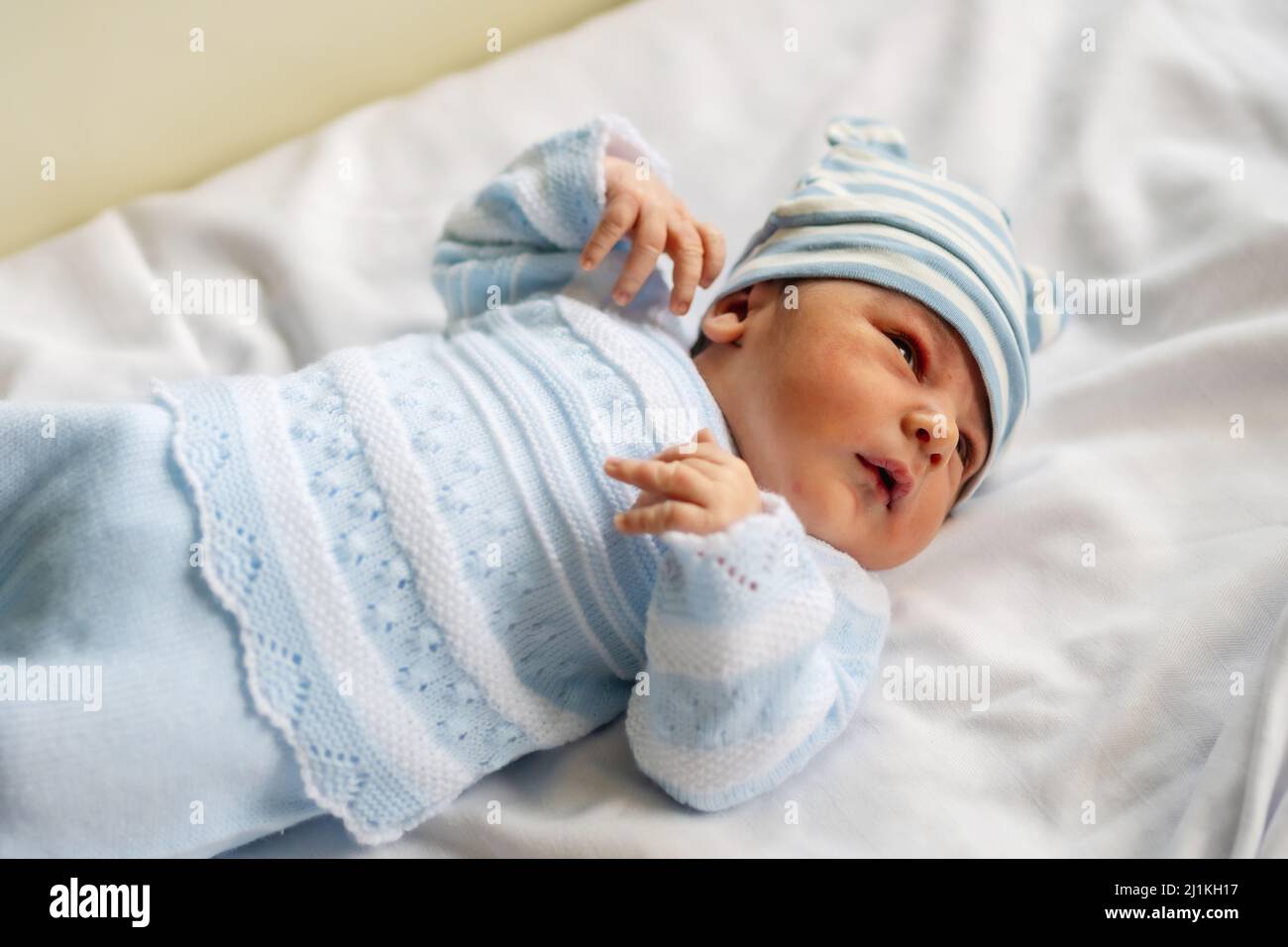 Una foto di un neonato in ospedale primo giorno di vita con una tuta bianca un cappello blu e sdraiato nella culla Foto stock Alamy