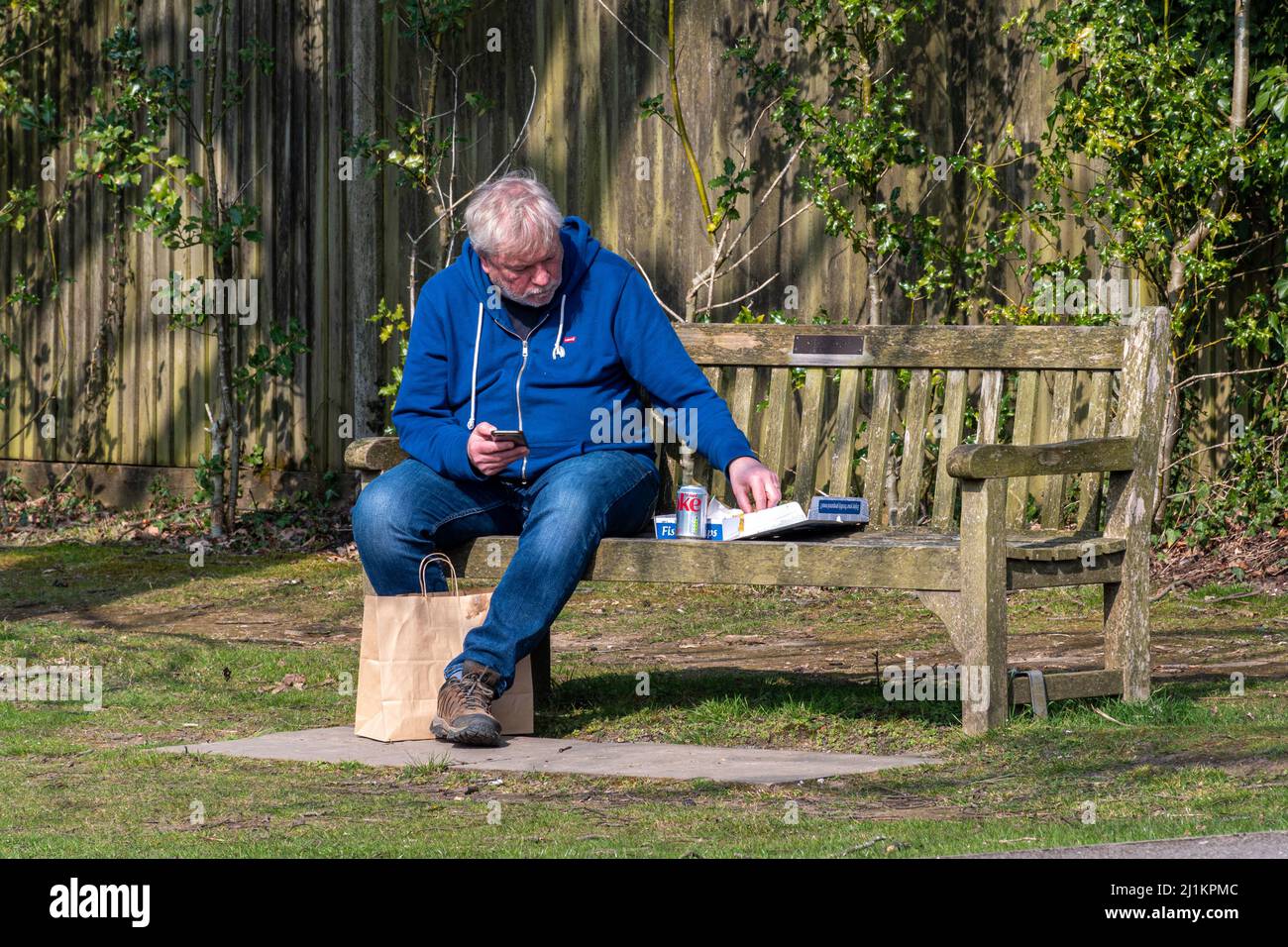 Uomo che mangia pesce da asporto e patatine pasto con una lattina di coca cola seduta su una panchina villaggio, Inghilterra, Regno Unito Foto Stock