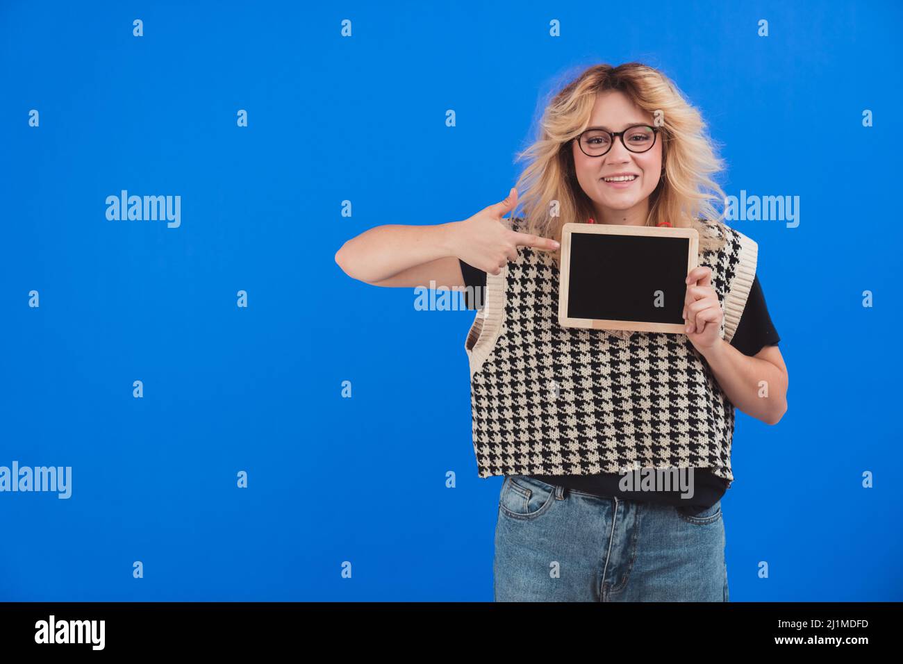Giovane donna bionda caucasica che tiene una tavoletta e punta con un dito indice. Spazio di copia. Sfondo blu isolato. Foto di alta qualità Foto Stock