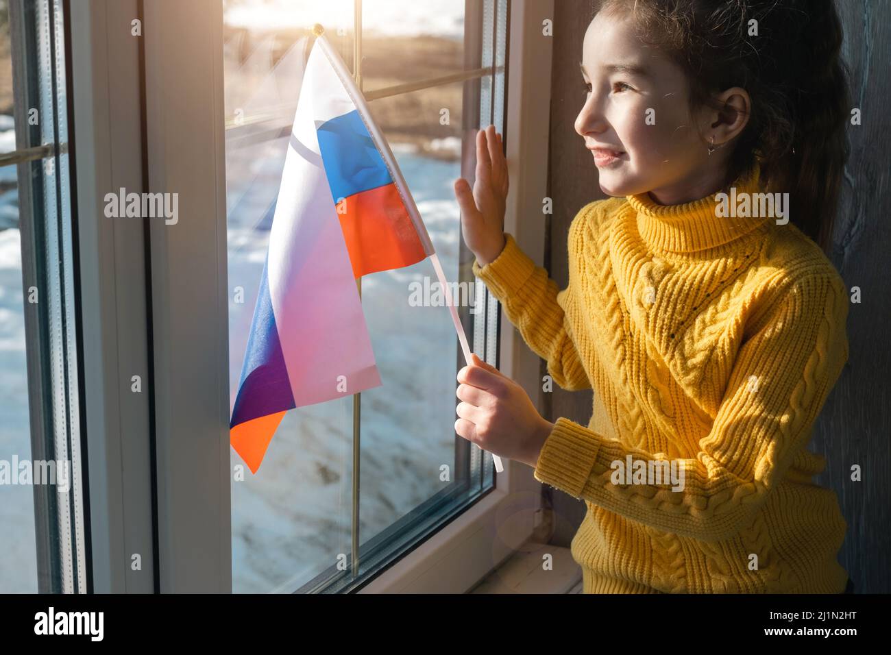 Una ragazza gioiosa in un maglione giallo con la bandiera della Russia guarda fuori la finestra. Patriot, pace nel mondo, Giornata della Vittoria, sostegno e sicurezza Foto Stock