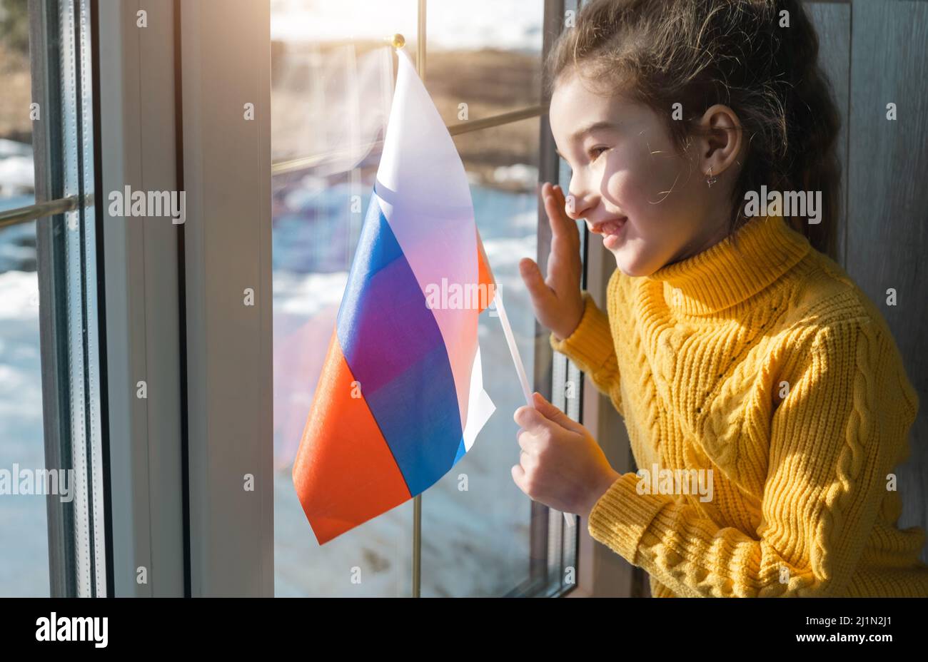 Una ragazza gioiosa in un maglione giallo con la bandiera della Russia guarda fuori la finestra. Patriot, pace nel mondo, Giornata della Vittoria, sostegno e sicurezza Foto Stock