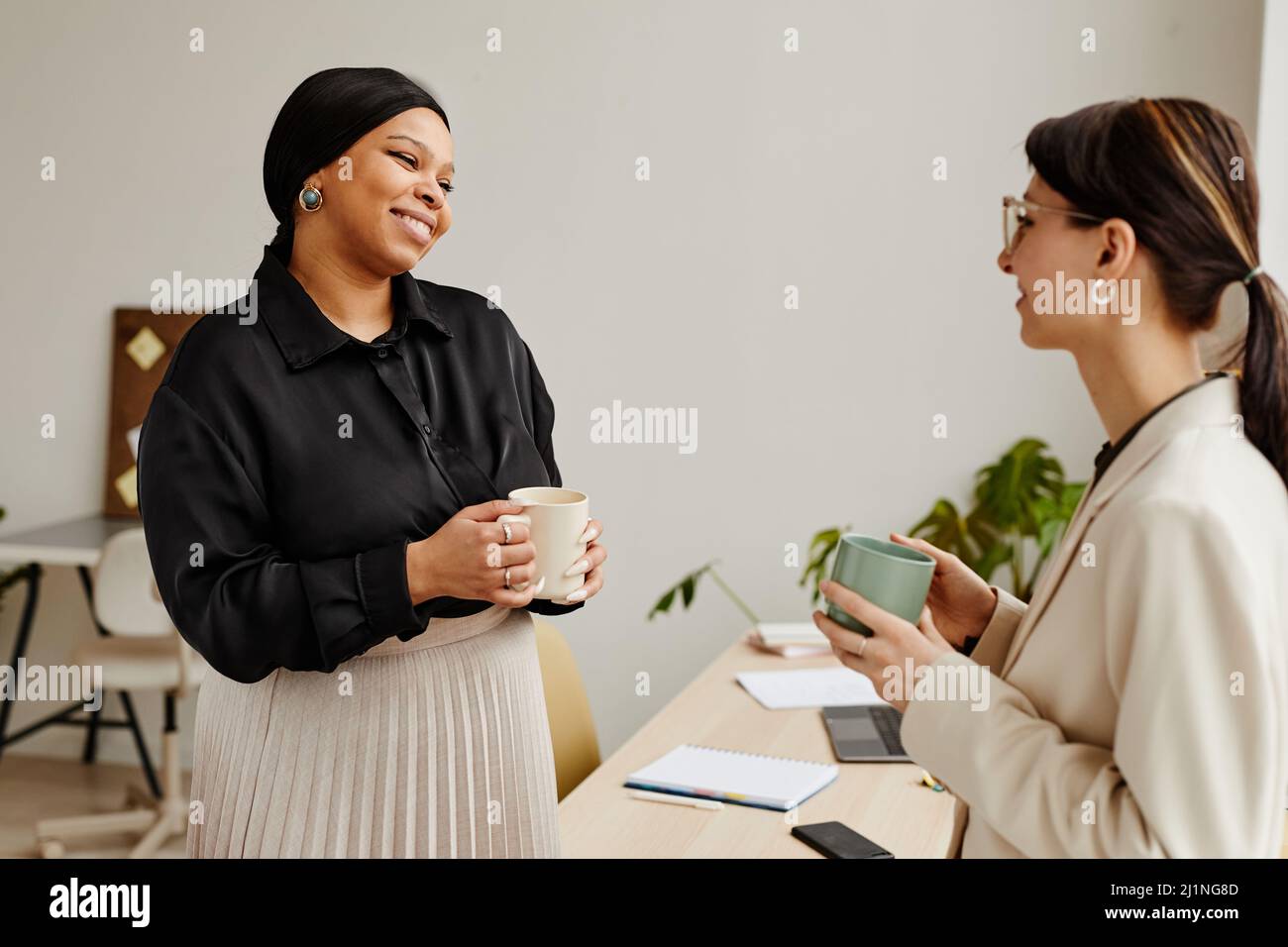 Ritratto minimo di due giovani donne di successo che chiacchierano alla pausa caffè in ufficio, fuoco sulla donna d'affari nera sorridente Foto Stock