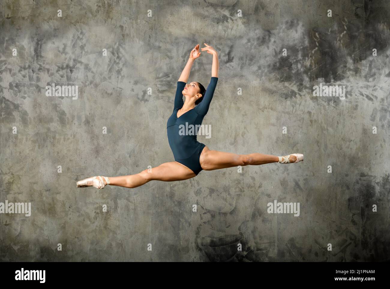 Corpo pieno di grazioso istruttore di balletto femminile in tuta e scarpe a punta che mostrano il classico salto gran jete contro il muro grigio di cemento Foto Stock