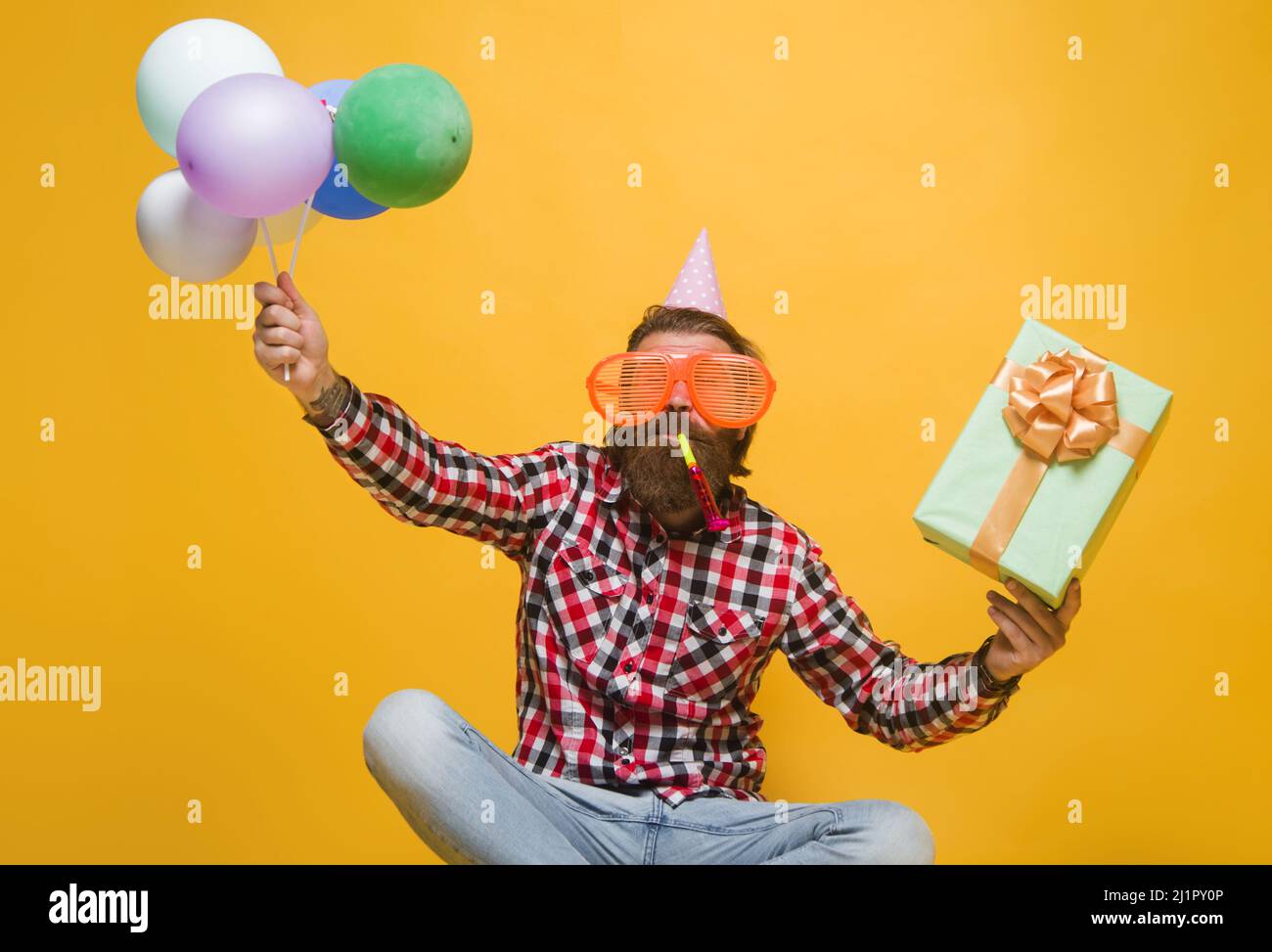 Ragazzo felice festeggiava il compleanno. L'uomo allegro barbuto in cappello di festa con i palloncini e la scatola attuale. Foto Stock