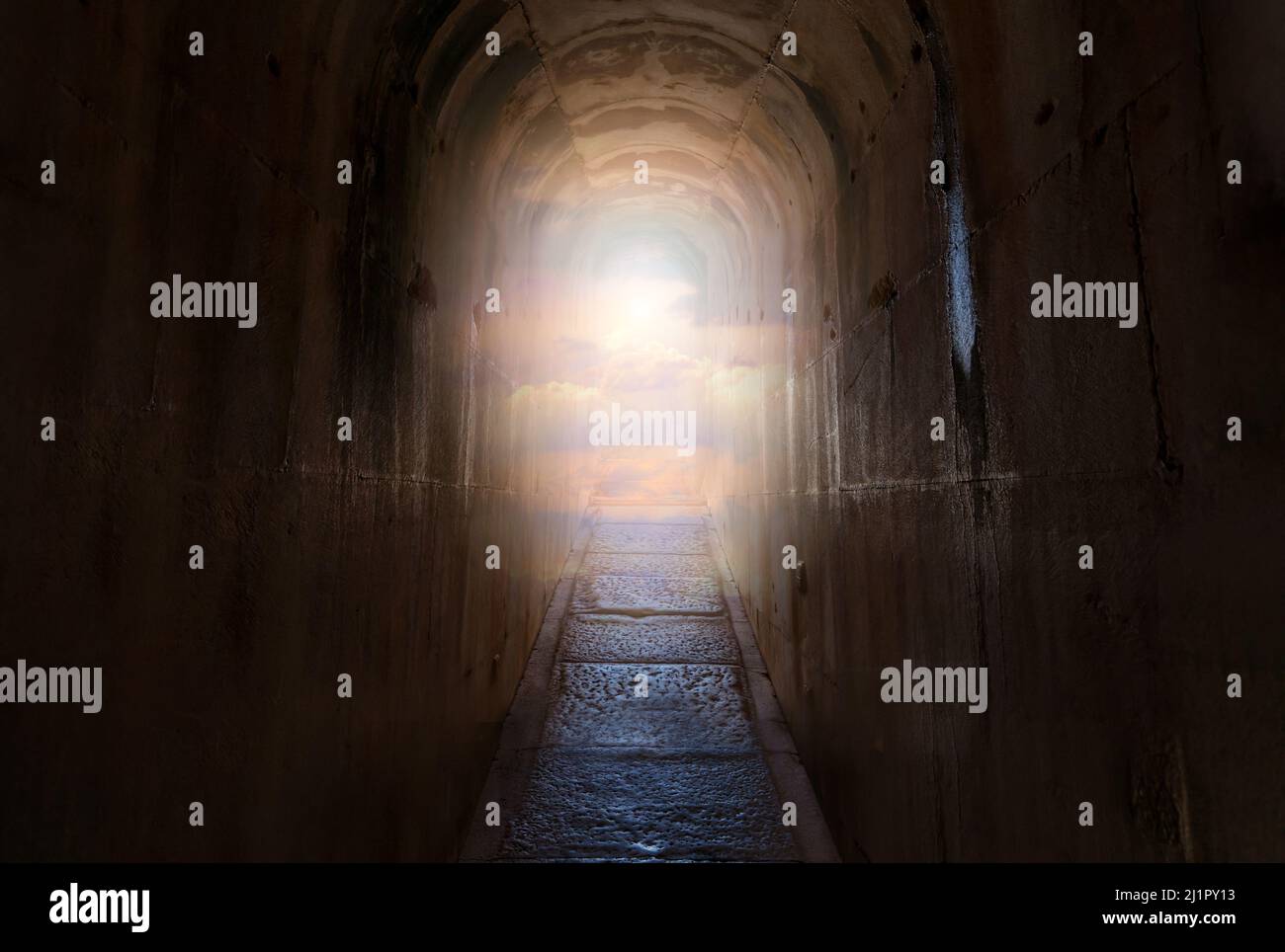 Tunnel scuro con sentiero stretto e fine del percorso che splende cielo e nuvole. Concetto di religione del paradiso o del paradiso. Foto Stock