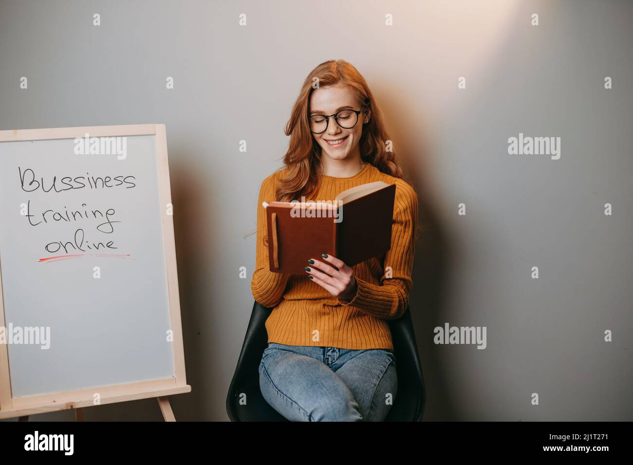 Donna che prende le lezioni in linea sul commercio seduto sulla sedia vicino ad una lavagna bianca e tenendo un libro nelle sue mani. Giornata felice di alfabetizzazione. Buon giorno dell'insegnante. Foto Stock