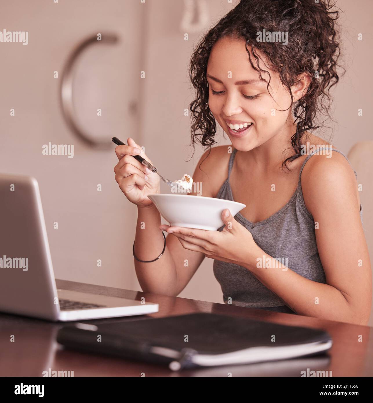 Sono sempre di umore migliore dopo colazione. Scatto di una donna che mangia la colazione mentre si siede al suo portatile. Foto Stock