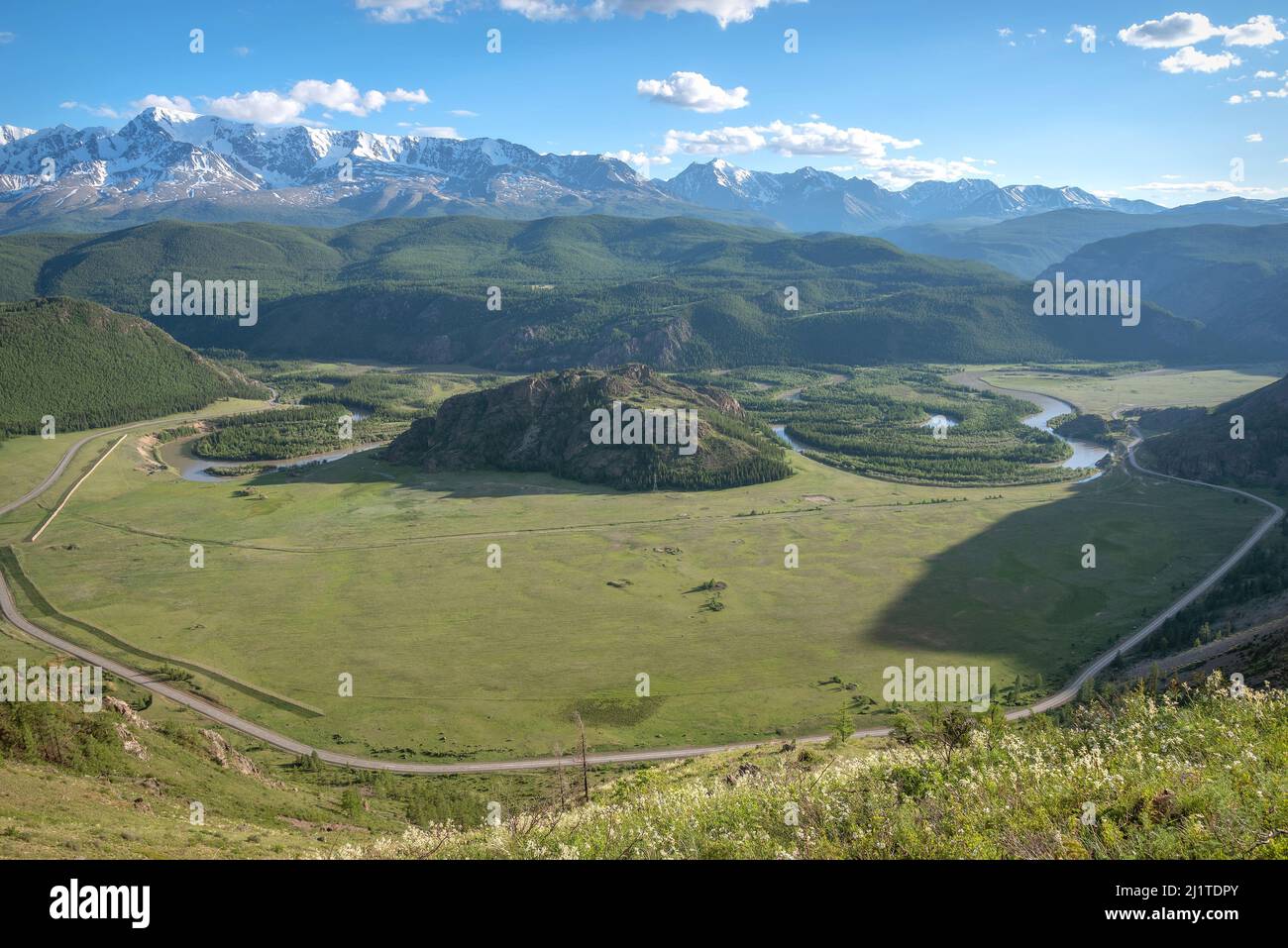 Bella vista aerea estiva di un fiume tortuoso, montagne innevate, una foresta sulle pendici delle montagne e una strada asfaltata contro un cielo blu con Foto Stock