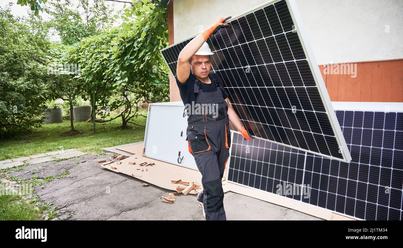 Uomo serio installatore solare che trasporta il modulo solare nel cortile. Lavoratore maschile in tute da lavoro con pannelli solari fotovoltaici e camminando lungo il percorso asfaltato fuori dalla casa. Foto Stock