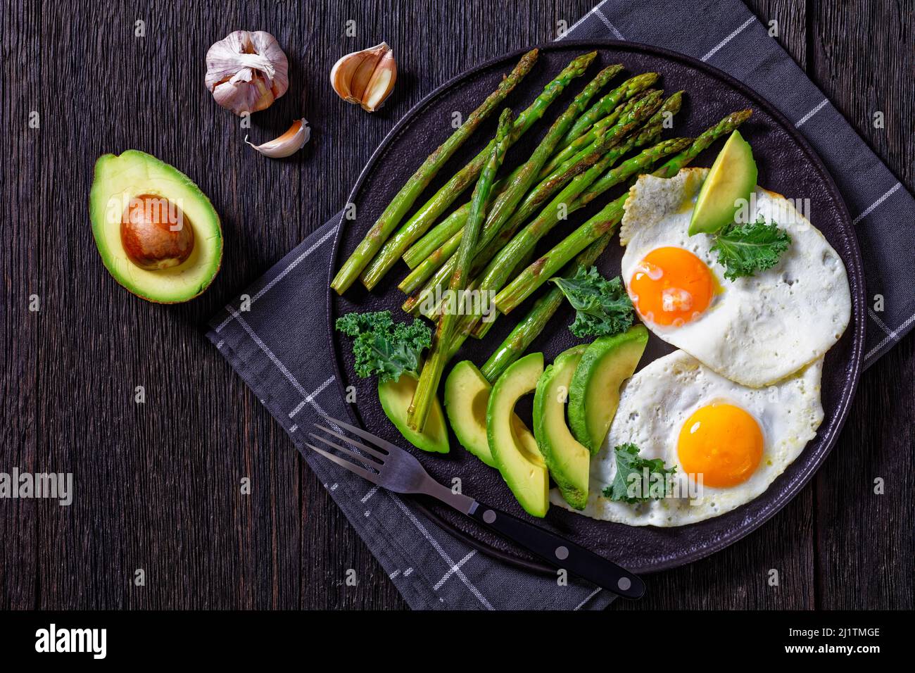asparagi arrostiti, avocado fresco maturo e uova fritte con tuorli runny su un piatto nero su un tavolo di legno scuro, piatto Foto Stock