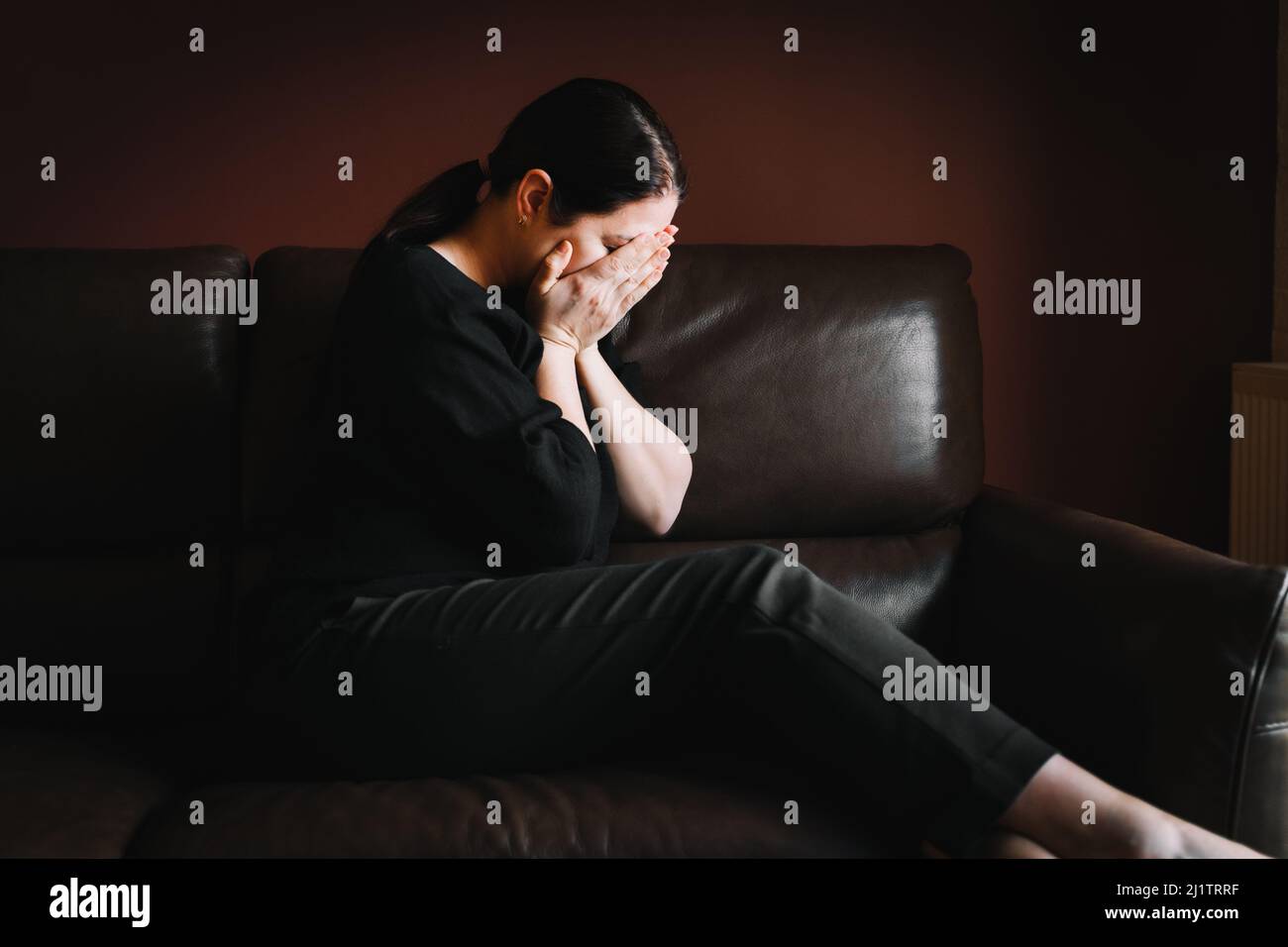 Giovane donna con capelli lunghi e scuri che indossa una blusa nera, pantaloni, seduta su un divano marrone, coprendo il viso con le mani. Foto Stock