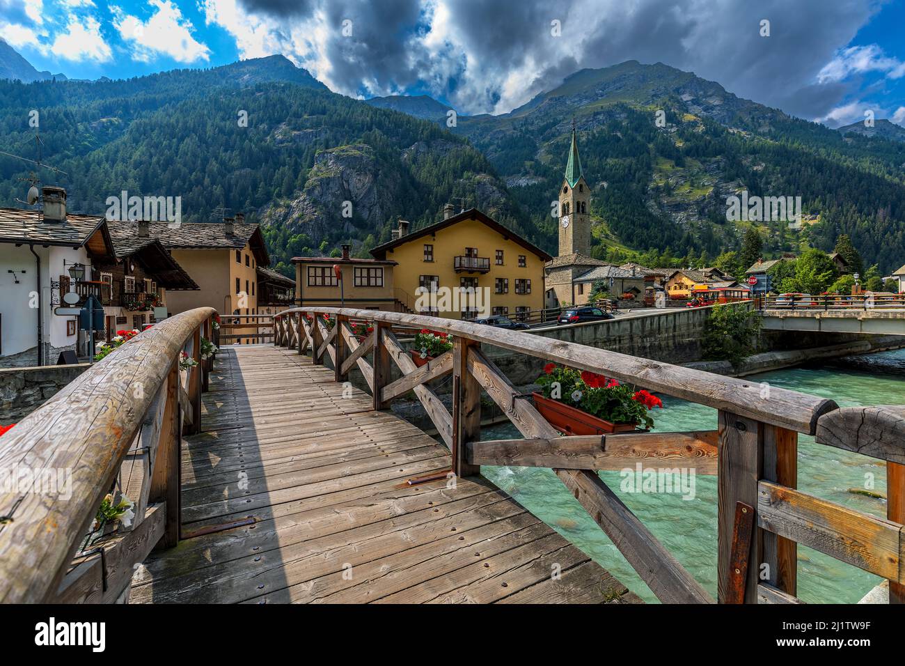 Ponte di legno sul fiume di montagna come case e vecchia chiesa sullo sfondo nel piccolo comune di Gressoney-Saint-Jean in Valle d'Aosta, Italia. Foto Stock