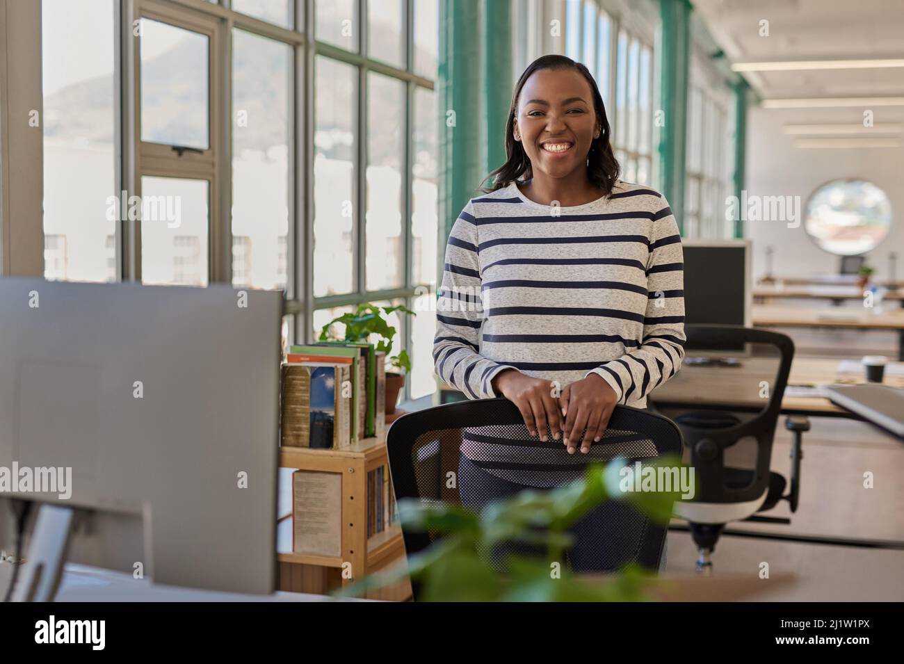 Sorridente giovane donna d'affari africana in piedi accanto alla sua scrivania in un ufficio Foto Stock