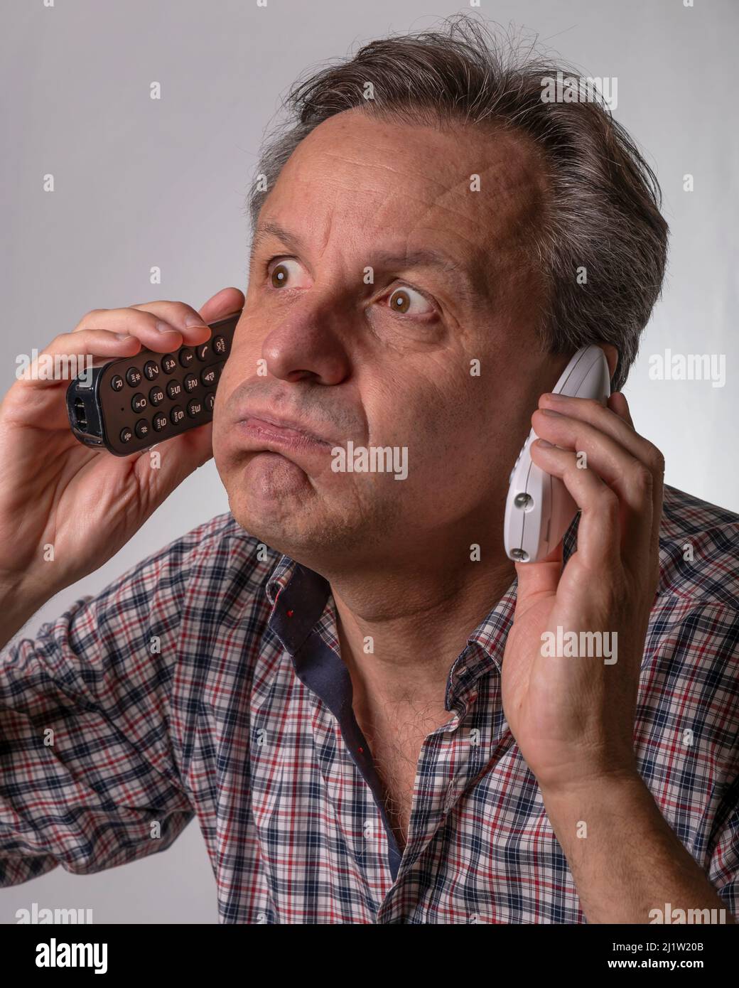 Un uomo con un'espressione scioccata ascolta due telefonate da due diversi telefoni cordless che ne tiene uno in ogni orecchio Foto Stock