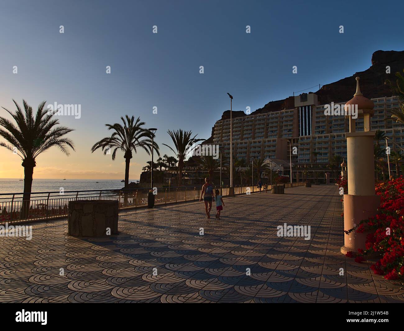 Persone che camminano sul lungomare della località turistica Taurito sulla costa meridionale di Gran Canaria, Spagna in serata luce del sole con palme. Foto Stock