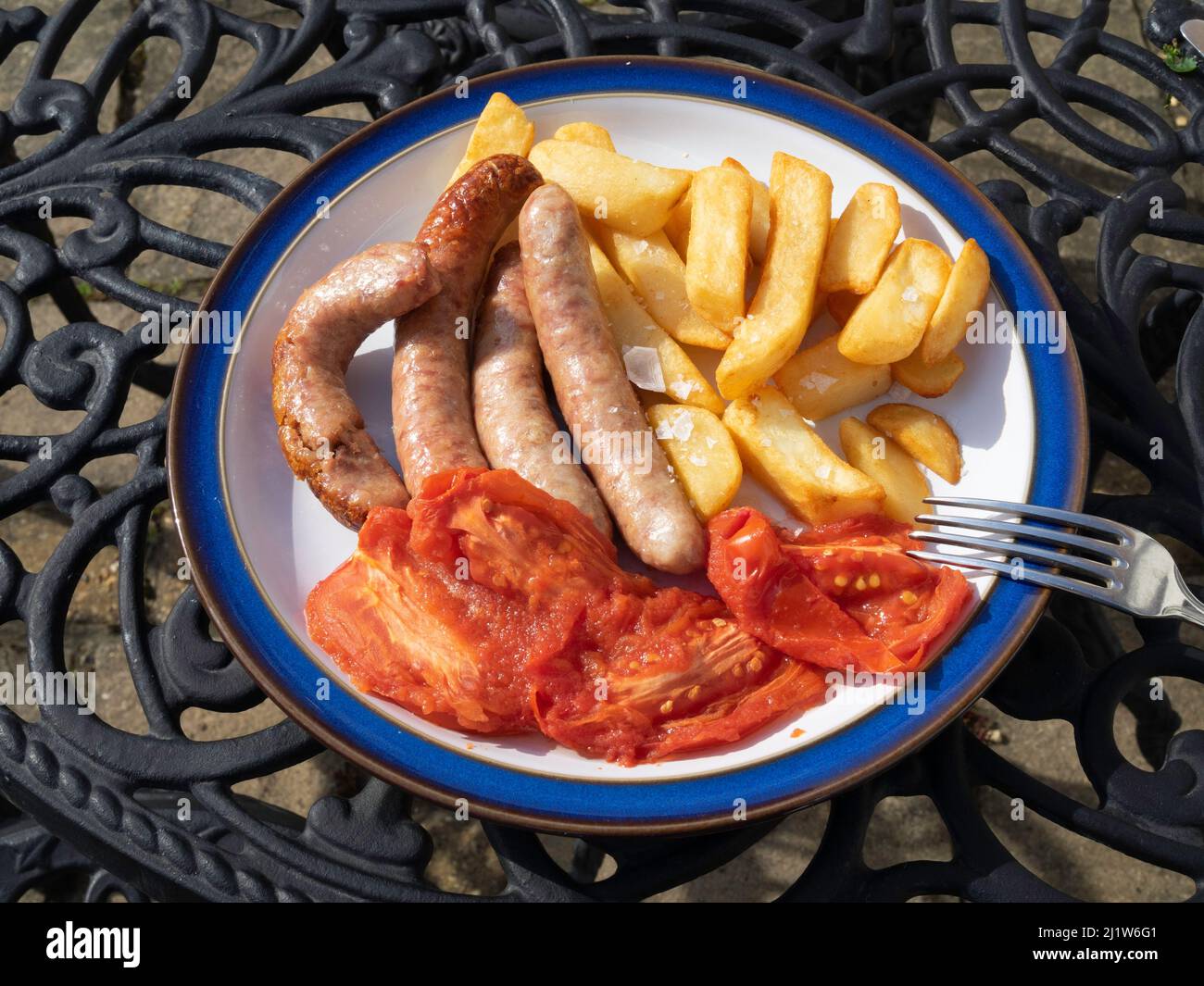Pranzo all'aperto con le tradizionali salsicce di maiale, pomodori prugna alla griglia e patatine fritte su un piatto bianco dal bordo blu Foto Stock