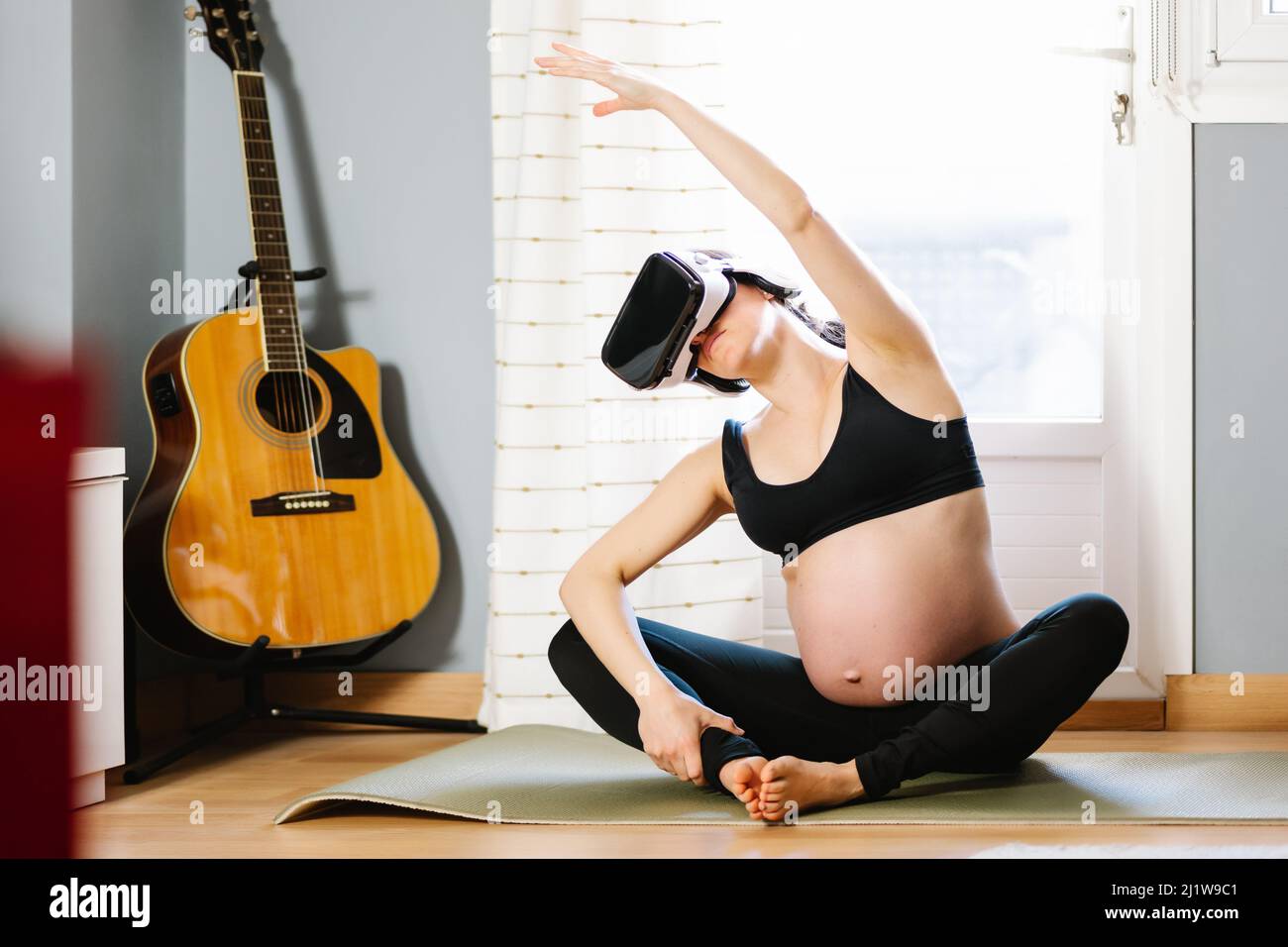 Corpo pieno di donna incinta allegra in abbigliamento sportivo e occhiali VR seduti in posa Butterfly e fare yoga nel cyberspazio in salotto Foto Stock