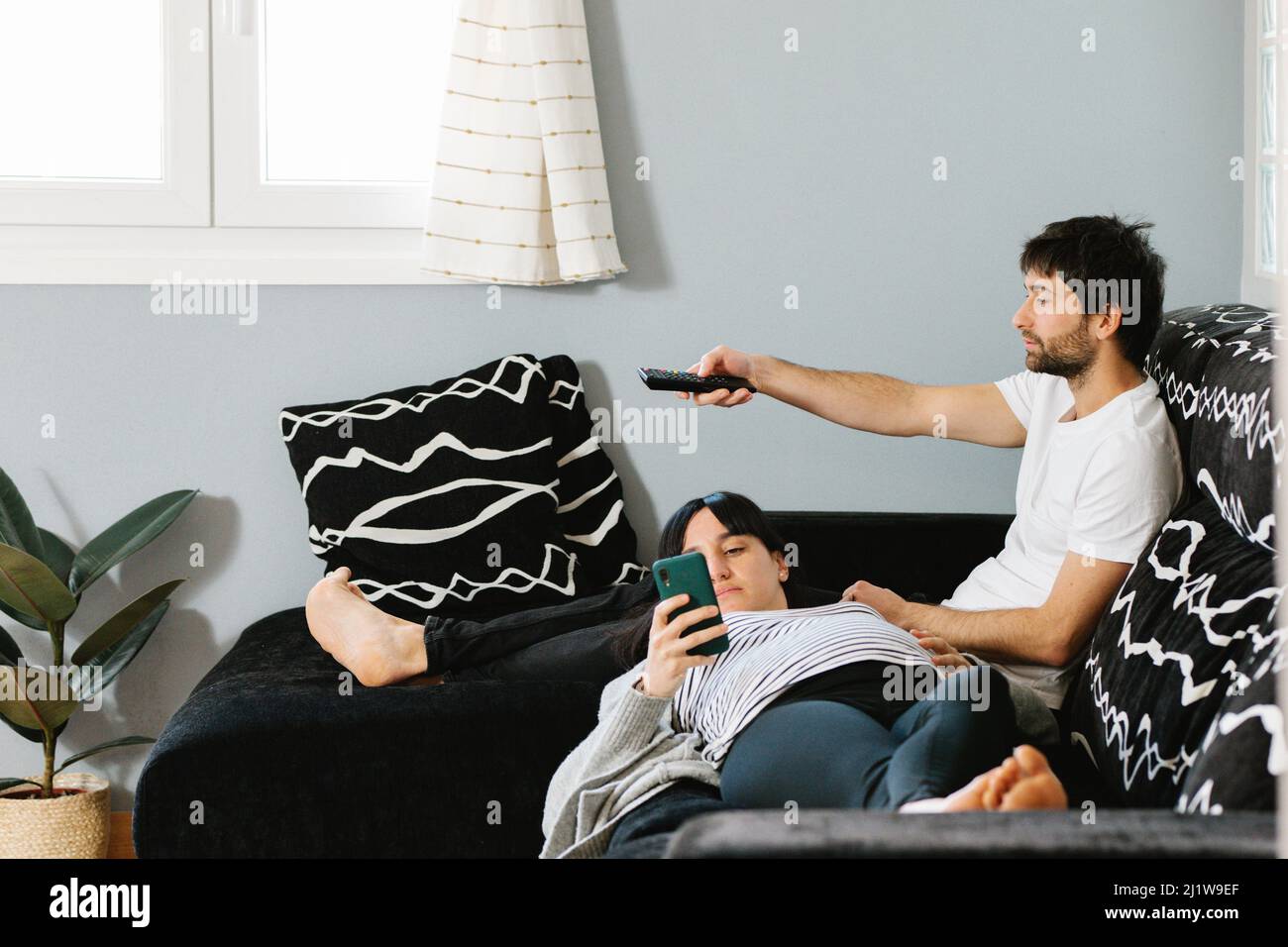 Corpo pieno di giovane etnia incinta barefooted signora usando il telefono mobile mentre si trova sulle ginocchia del marito seduto sul divano e i canali di commutazione di dur della TV Foto Stock