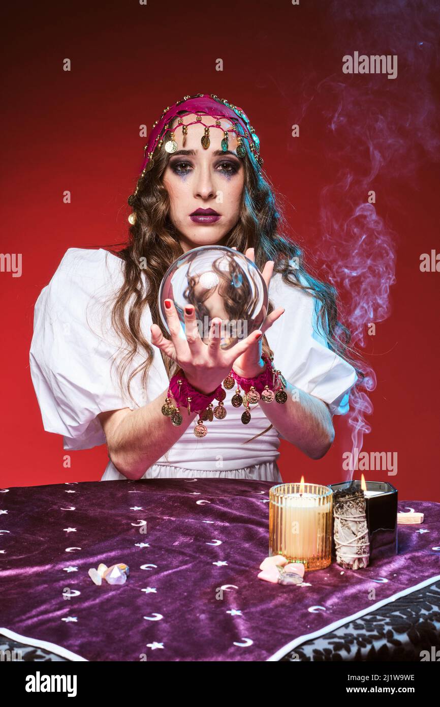 Affascinante donna fortuna teller con trucco indovinare futuro a tavola con palla magica e candele bruciando vicino al cristallo Foto Stock