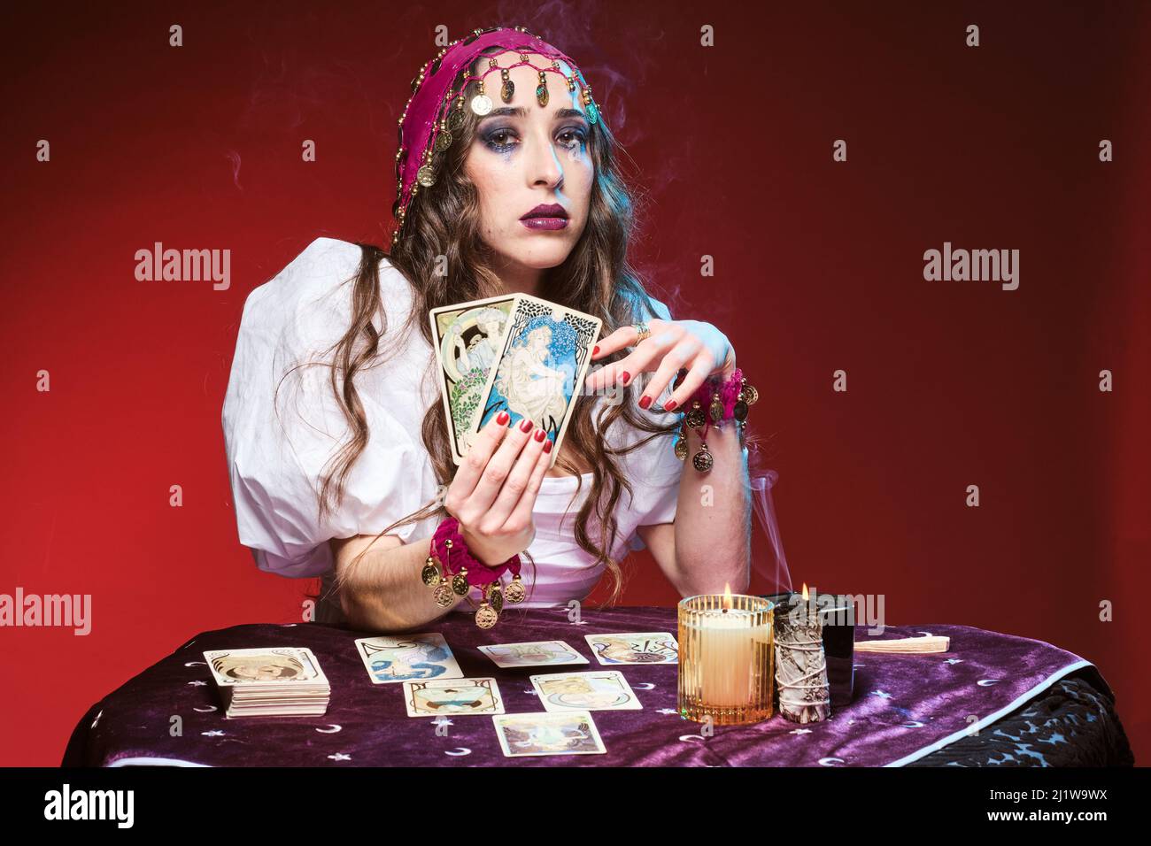 Attraente soothsayer femminile con trucco seduto al tavolo con carte tarocchi in mano contro candele brucianti e guardando la macchina fotografica Foto Stock