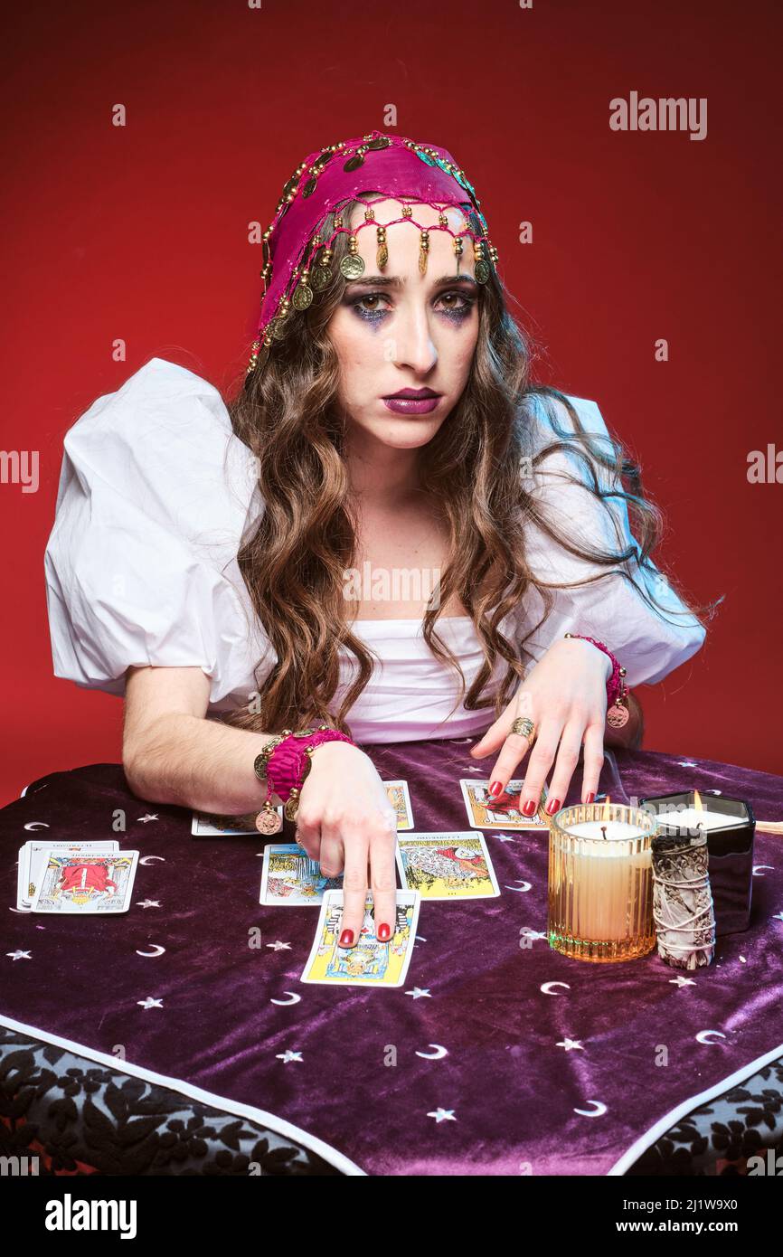 Attraente soothsayer femminile con trucco seduto al tavolo con carte tarocchi contro candele brucianti e guardando la macchina fotografica Foto Stock