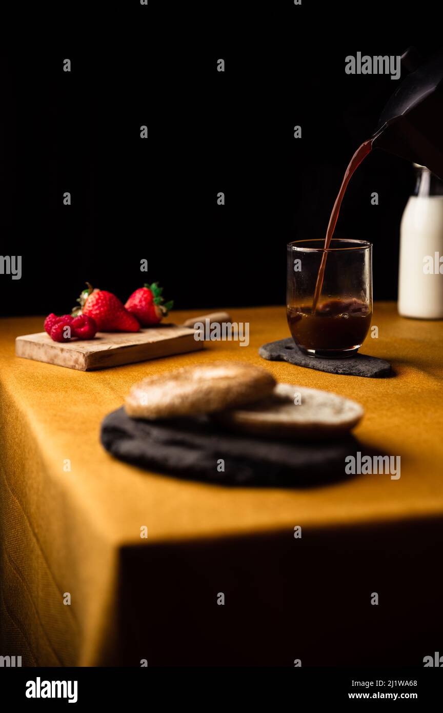 Caffè fresco e fragrante che si versa in un bicchiere su un piatto di ardesia dalla moka pot per colazione con bagel e lamponi con fragole Foto Stock