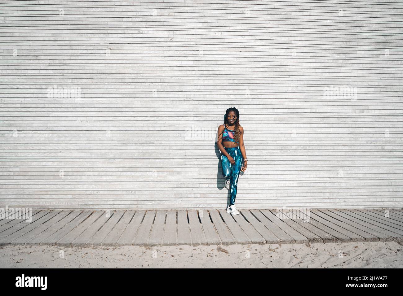 Corpo pieno di femmina afroamericana sorridente sottile con lunghi dreadlock in reggiseno sportivo e leggings in piedi su passerella di legno e appoggiati su parete di metallo Foto Stock
