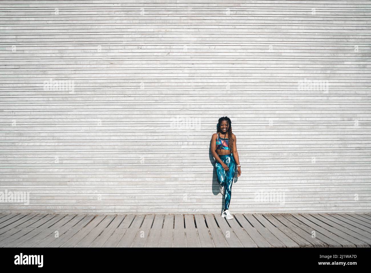 Corpo pieno di femmina afroamericana sorridente sottile con lunghi dreadlock in reggiseno sportivo e leggings in piedi su passerella di legno e appoggiati su parete di metallo Foto Stock