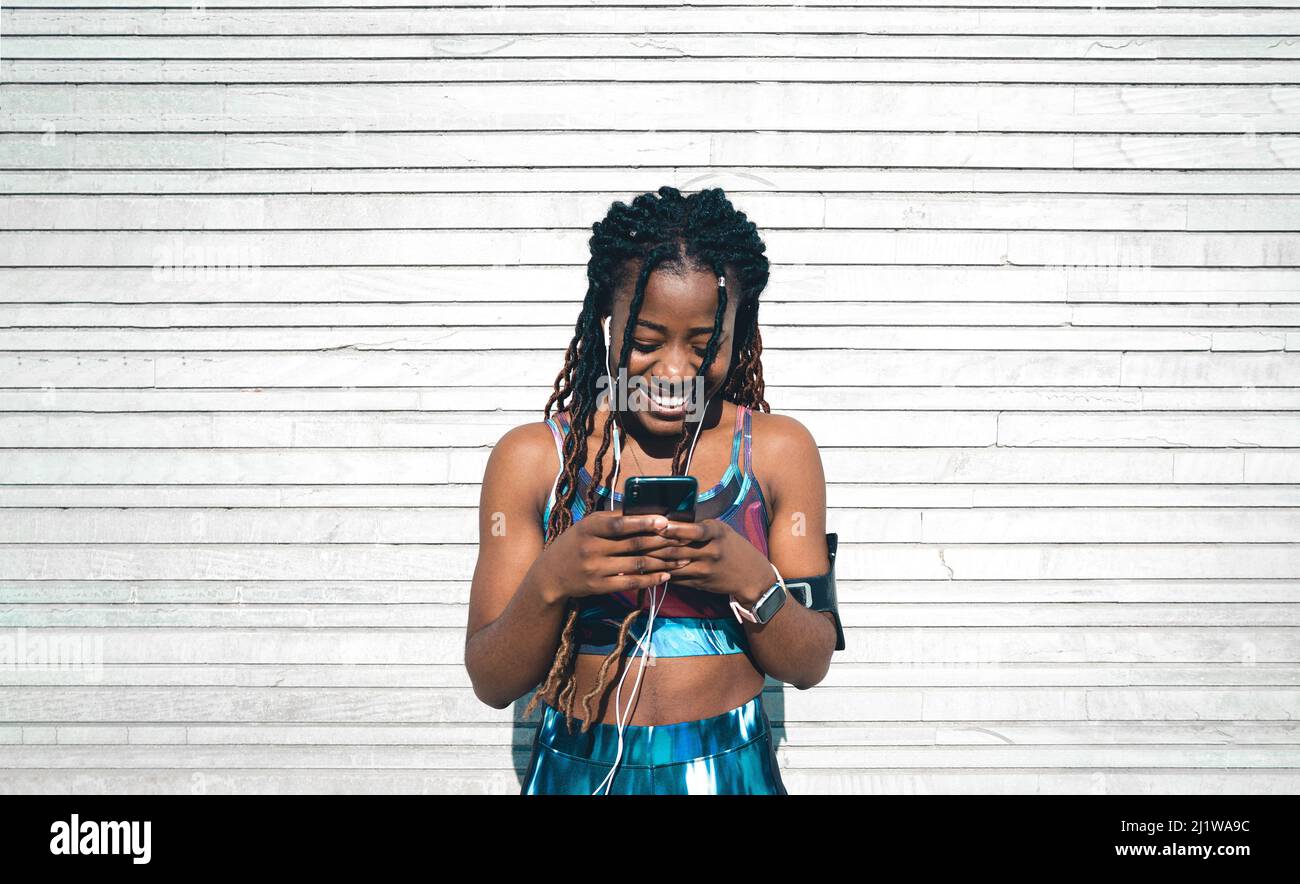 Vista frontale della giovane ragazza afro-americana felice con dreadlock indossando abbigliamento sportivo ascoltare musica in auricolari e navigare cellulare mentre magro Foto Stock