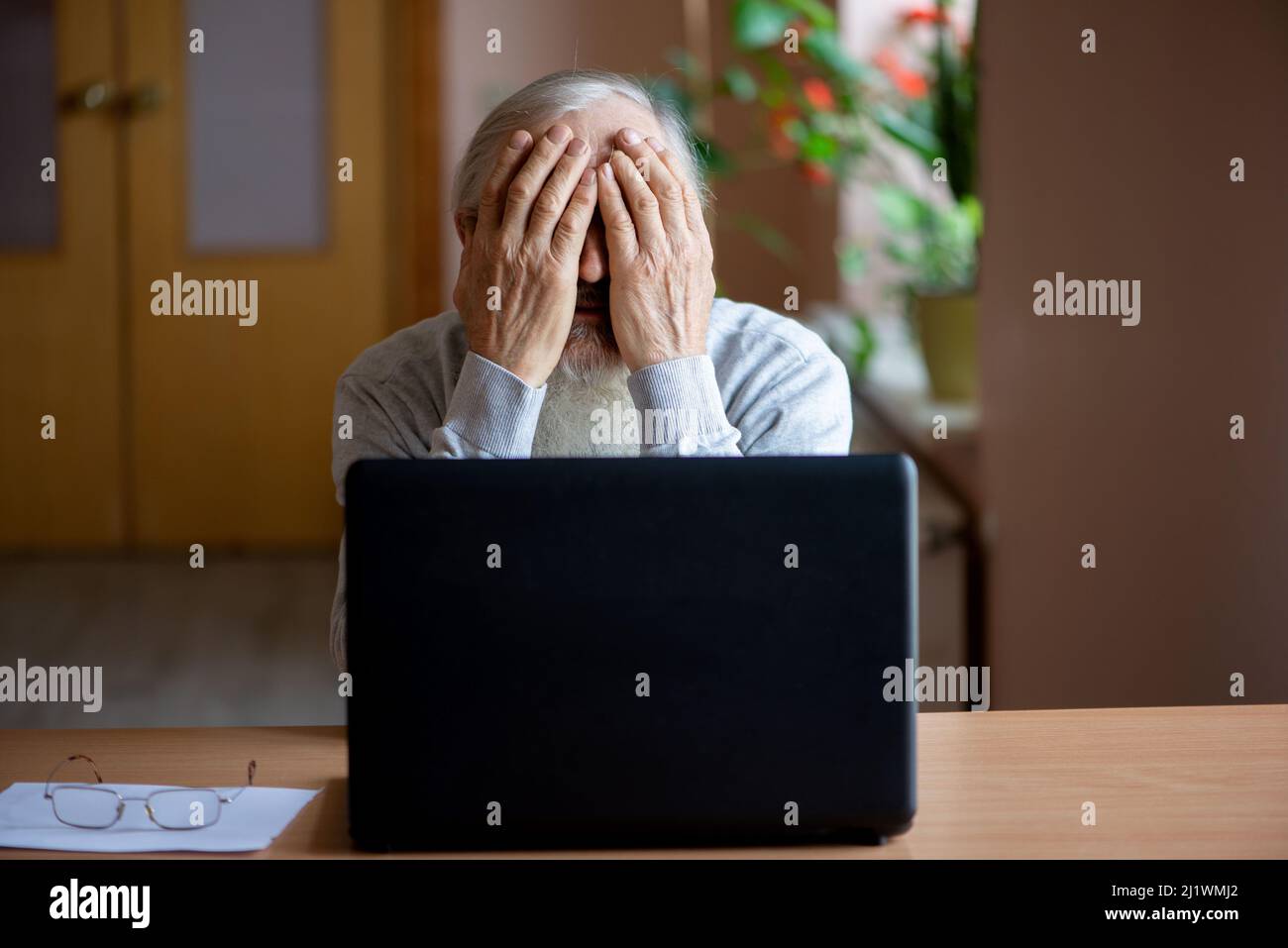 Un anziano stanco, rotto, rassegnato, preoccupato si siede davanti ad un computer portatile e gli copre il volto con la mano. Il triste anziano controlla il suo ac Foto Stock