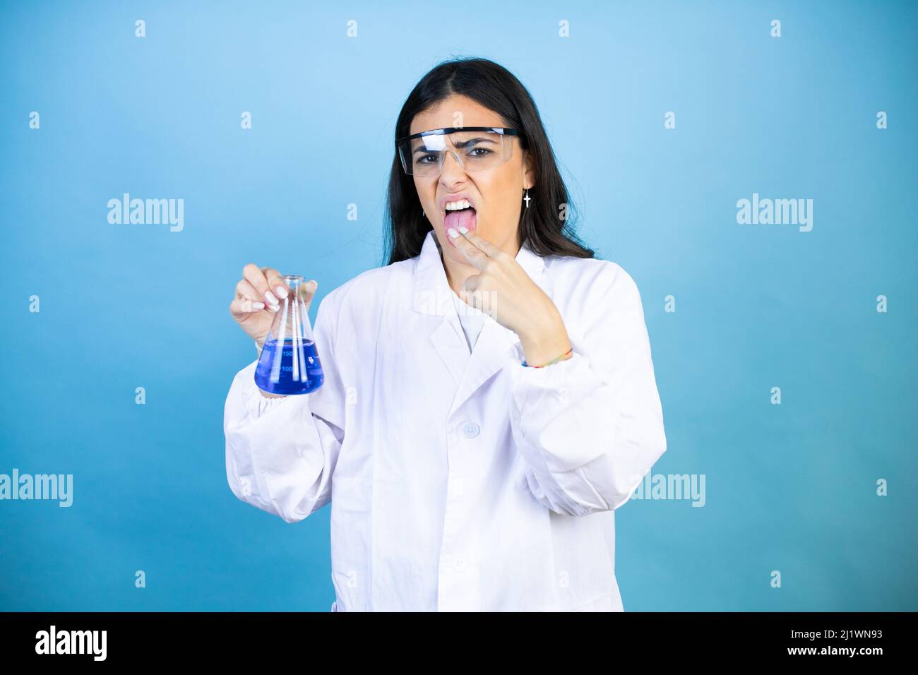 Giovane donna bruna indossando scienziato uniforme tenendo provetta su fondo blu isolato travestito con la mano all'interno della bocca Foto Stock