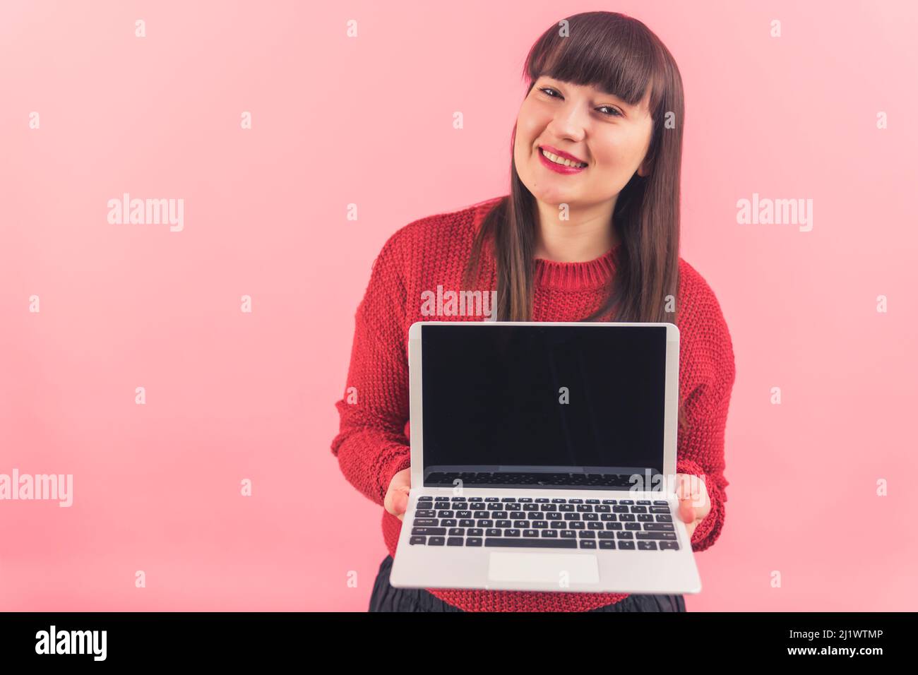 Sorridente bella donna caucasica che mostra notebook con nero schermo mock-up alla fotocamera su sfondo rosa. Foto di alta qualità Foto Stock