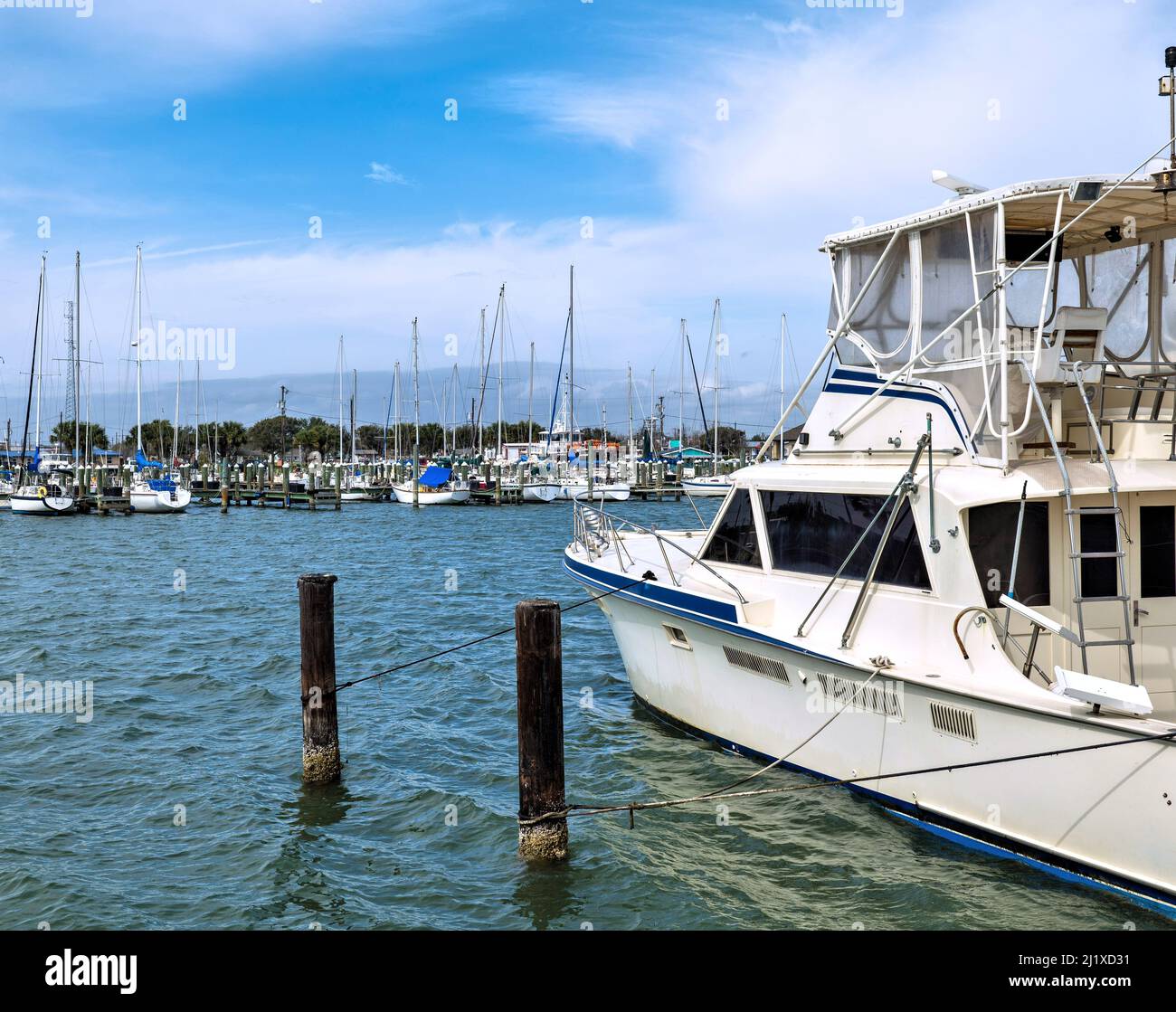 La barca a motore bianca da diporto è legata da corde a colonne di legno in acqua in un porticciolo con molti barche a vela in lontananza. Foto Stock