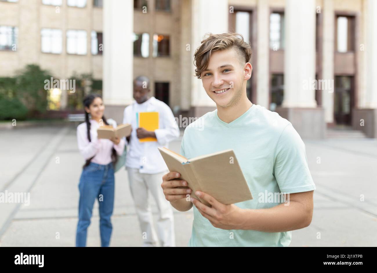 Corsi di laurea all'estero. Ritratto di felice studente maschio con libro in posa all'aperto al campus con i suoi compagni di classe Foto Stock
