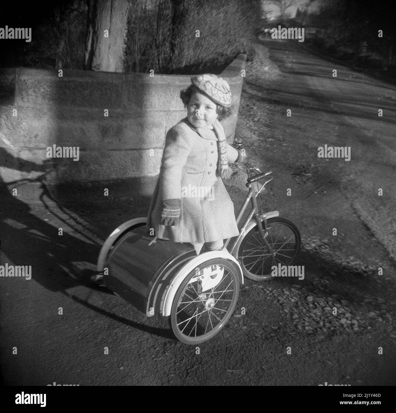 1950s, storica, fuori su un sentiero, una bambina dolce in un cappotto e cappello, seduto sul suo triciclo con stivale in metallo, Inghilterra, Regno Unito. Questo ciclo per bambini a tre ruote è stato realizzato da Triang, un produttore britannico leader di giocattoli per bambini a base di metallo in quest'epoca. Foto Stock