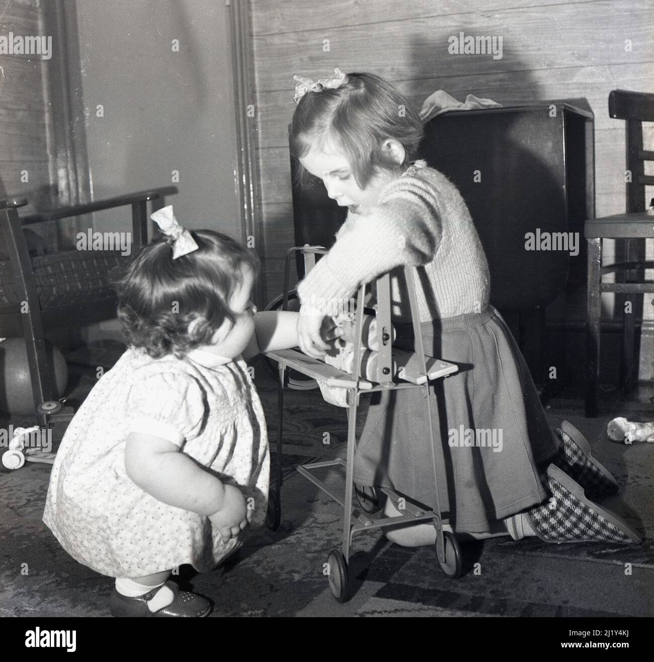 1950s, storica, una bambina e il suo piccolo assitore che gioca all'interno di una stanza con un giocattolo mangle, Inghilterra, Regno Unito. Il mangle giocattolo con telaio in metallo, con rulli in legno e su ruote è stato fatto da Triang, un produttore britannico leader di giocattoli a base di metallo per bambini in questa epoca. Le mangles o stringers come erano anche conosciuti, erano un articolo domestico usato nella casa in questa ed era precedente nel lavoro di lavanderia, per spremere o strizzare l'acqua dai vestiti lavati prima di asciugare. Foto Stock