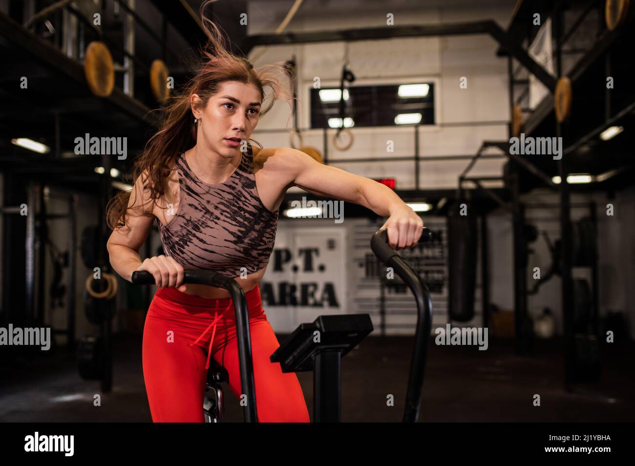 Una giovane donna muscolare attraente esercita duro su una bicicletta nel suo abbigliamento sportivo e sudore. Allenamento individuale in una palestra moderna e completamente attrezzata. Foto Stock