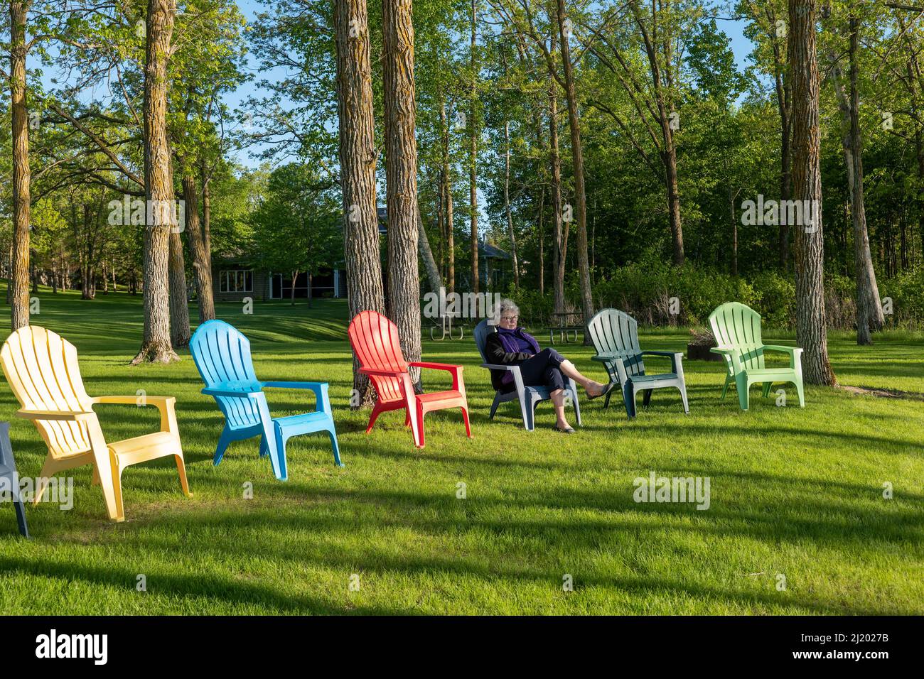 Una donna seduta su una sedia Adirondack con alberi e boschi sullo sfondo. Ombre. Foto Stock