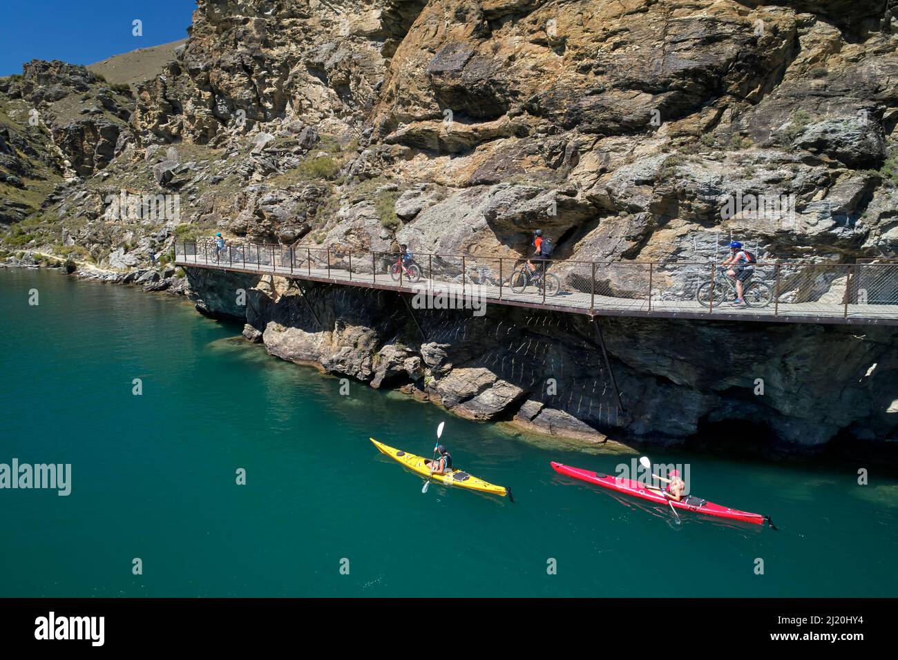 Ciclisti sul ponte a sbalzo sul percorso ciclistico del lago Dunstan e kayak sul lago Dunstan, vicino a Cromwell, Otago centrale, Isola del Sud, Nuova Zelanda - drone Foto Stock