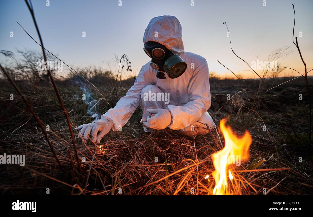Persona irriconoscibile che brucia vecchia erba secca in campo. Ecologo in maschera di gas che tiene matchbox e la messa a fuoco per asciugare erba. Ricercatore scienziato che indossa tuta protettiva e respiratore. Foto Stock